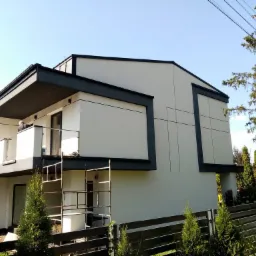 Nowoczesny, dwupiętrowy dom z płaskim dachem, wykończony w bieli i szarości, z widocznymi rusztowaniami przy balkonie i elementami dekoracyjnymi na elewacji, otoczony zielenią i ogrodzeniem.