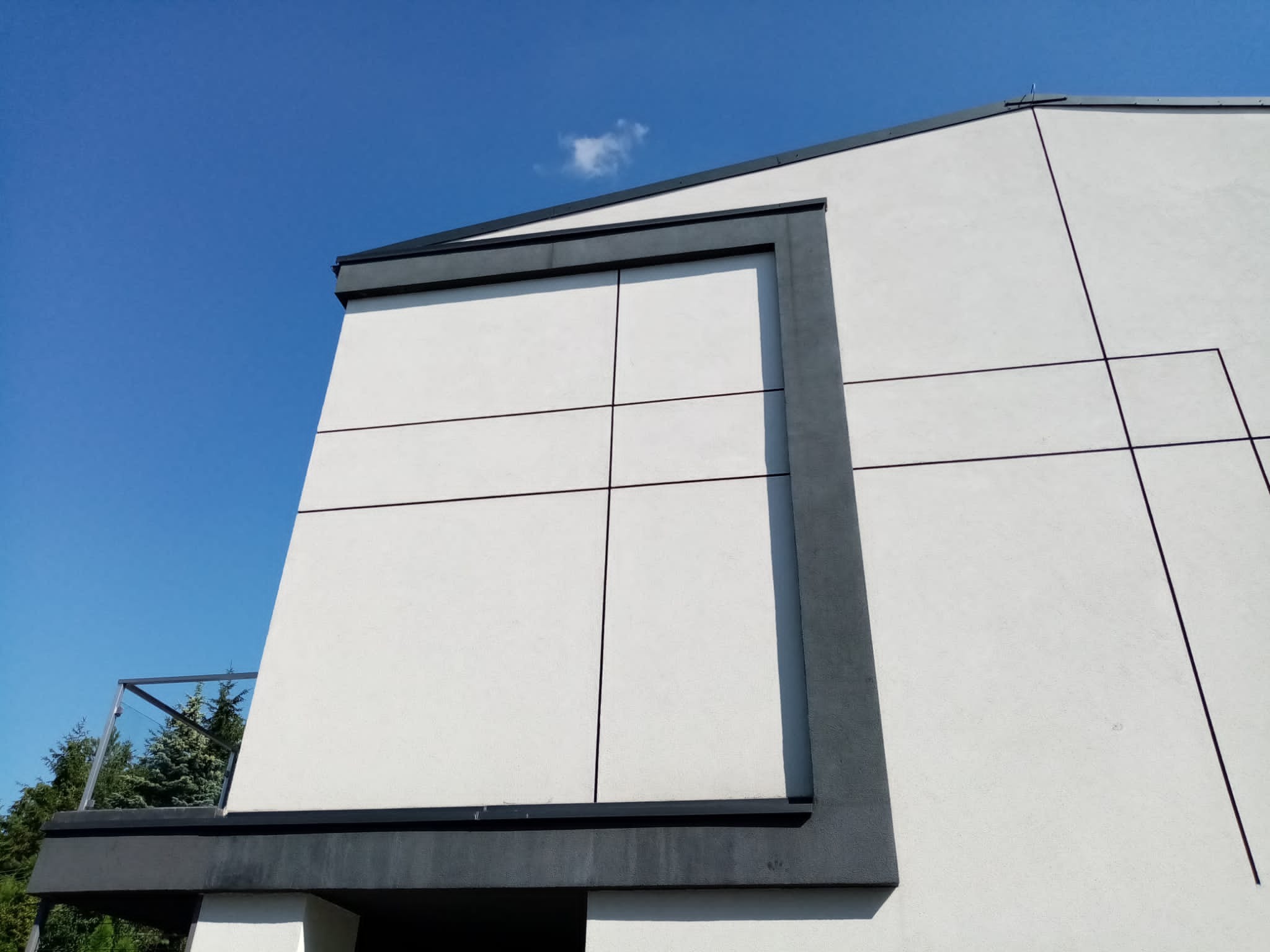 Fragment elewacji budynku z białym tynkiem i szarymi elementami dekoracyjnymi, widoczne pionowe i poziome podziały, na górze fragment dachu, w tle błękitne niebo.