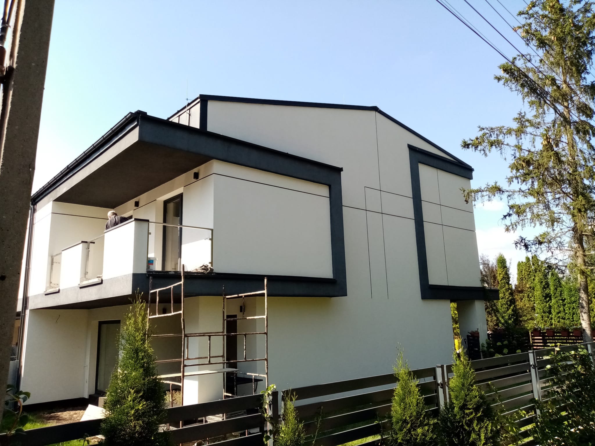 Nowoczesny, dwupiętrowy dom z płaskim dachem, wykończony w bieli i szarości, z widocznymi rusztowaniami przy balkonie i elementami dekoracyjnymi na elewacji, otoczony zielenią i ogrodzeniem.