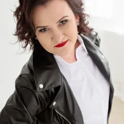 Portret kobiety w czarnej skórzanej kurtce i białej koszuli, z intensywnie czerwoną szminką, patrzącej w górę w obiektyw