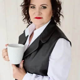 Kobieta w białej koszuli i czarnej kamizelce trzyma biały kubek z wypukłą fakturą, na jasnym, drewnianym tle.