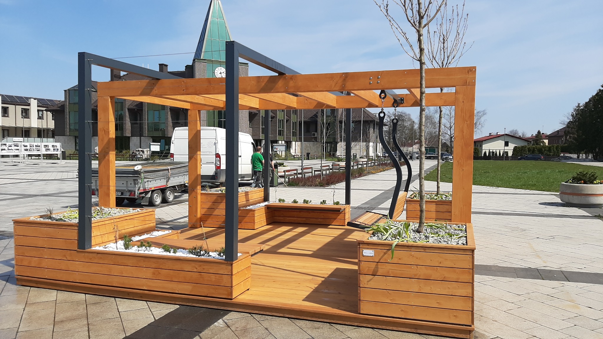 Drewniana pergola z metalowymi elementami, zintegrowana z donicami i siedziskami, na placu miejskim z kostki brukowej w słoneczny dzień. W tle budynki użyteczności publicznej i pracownik...