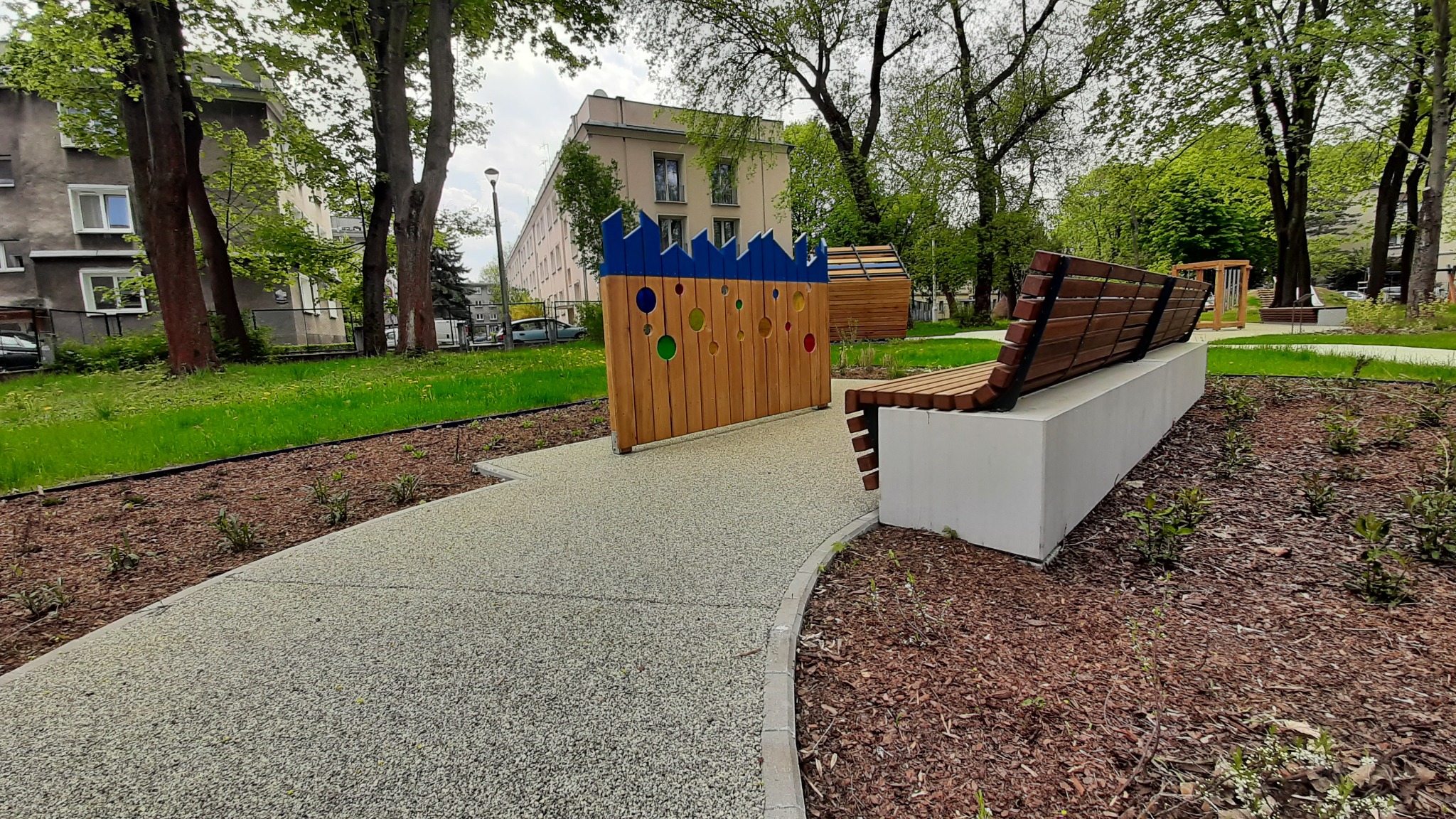 Ścieżka z nawierzchni mineralnej w parku miejskim, obok drewniana ławka na betonowym postumencie i ażurowa ścianka z kolorowymi otworami, w tle budynek mieszkalny i drzewa.