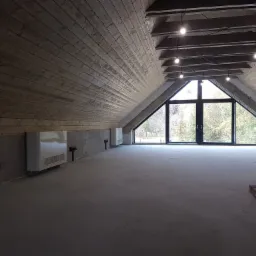 Poddasze z drewnianą boazerią na ścianach i suficie, grzejniki elektryczne zamontowane na ścianie, duże okno z widokiem na zewnątrz, surowa podłoga i oświetlenie punktowe.