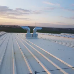 Widok z góry na dwa metalowe kominy wentylacyjne na białym dachu przemysłowym z widokiem na pola i zachodzące słońce.