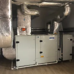 Komfovent Verso rekuperator z srebrnymi, izolowanymi przewodami wentylacyjnymi pod sufitem, widoczne rury doprowadzające media.