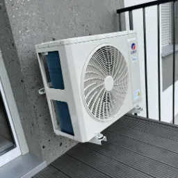 Biała jednostka zewnętrzna klimatyzacji marki Gree zamontowana na balkonie z szarą, teksturowaną ścianą i czarną balustradą, widoczna z boku.