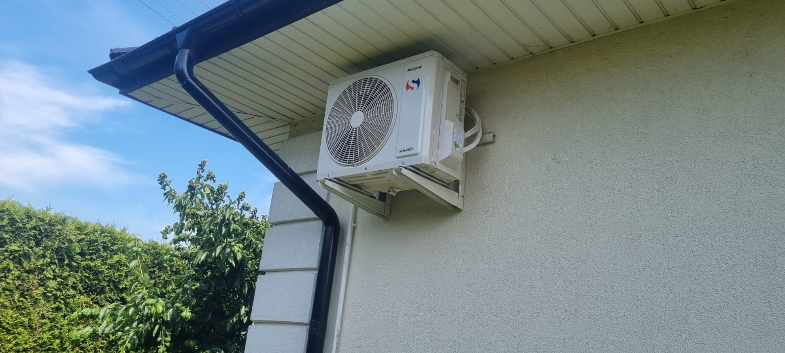 Biała jednostka zewnętrzna klimatyzacji zamontowana na elewacji domu, widoczny fragment rynny i zieleń w tle.