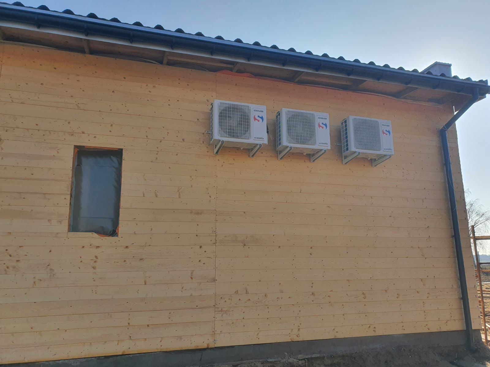 Trzy białe klimatyzatory zamontowane na zewnętrznej drewnianej ścianie domu w trakcie budowy, z oknem zabezpieczonym folią.