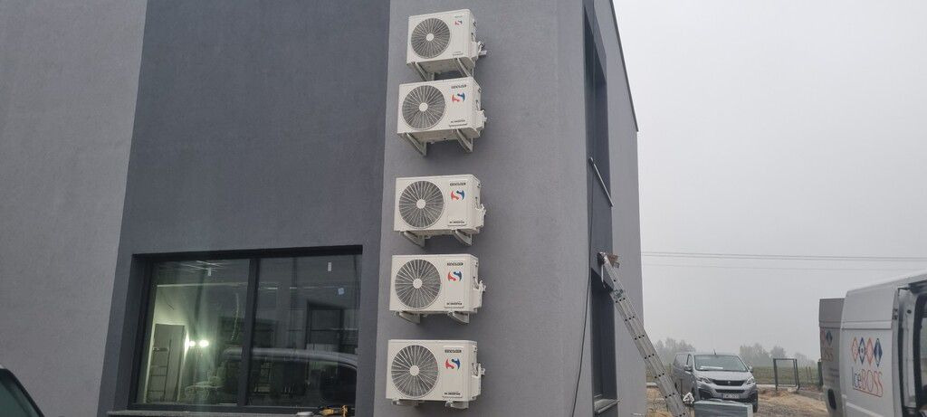 Rząd pięciu klimatyzatorów zamontowanych na szarej ścianie budynku, widok z dołu, drabina oparta o ścianę, samochody dostawcze w tle.