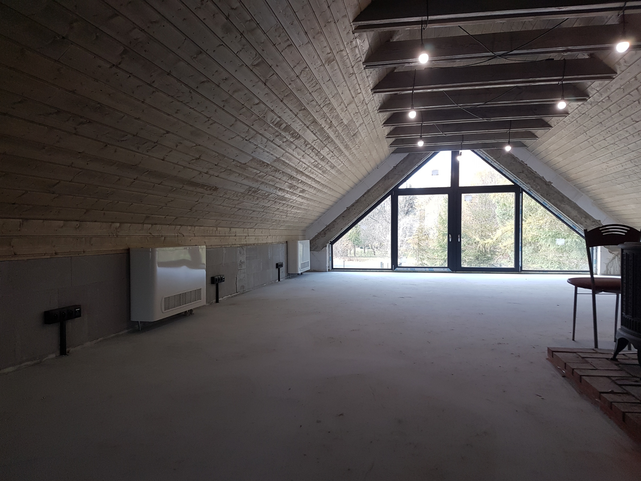 Poddasze z drewnianą boazerią na ścianach i suficie, grzejniki elektryczne zamontowane na ścianie, duże okno z widokiem na zewnątrz, surowa podłoga i oświetlenie punktowe.