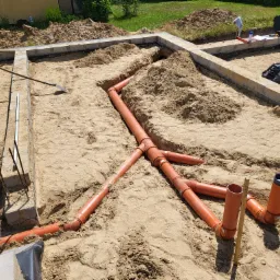 Pomarańczowe rury kanalizacyjne ułożone w wykopie fundamentowym na piaszczystym podłożu, widoczne narzędzia i elementy konstrukcyjne.