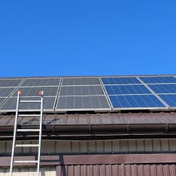 CLINER Marcin Dudziński - Panele słoneczne na brązowym dachu z blachy trapezowej, drabina oparta o ścianę budynku z kolcami przeciw ptakom, błękitne niebo w tle.