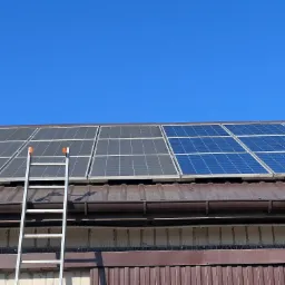 Panele słoneczne na brązowym dachu z blachy trapezowej, drabina oparta o ścianę budynku z kolcami przeciw ptakom, błękitne niebo w tle.