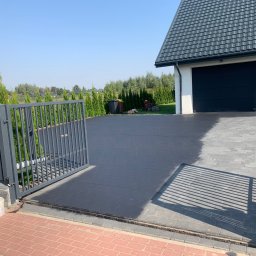 CLINER Marcin Dudziński - Ciemnoszara kostka brukowa na podjeździe do domu jednorodzinnego, widoczna brama wjazdowa, fragment elewacji z numerem 19 oraz garaż w tle, ślady po czyszczeniu kostki.