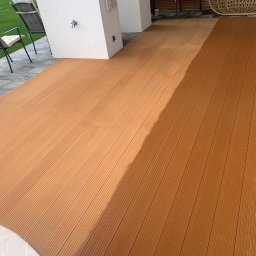 CLINER Marcin Dudziński - Częściowo umyta deska tarasowa z kompozytu w kolorze brązowym, widoczne różnice w odcieniach po czyszczeniu, na tle krzesła ogrodowe i fragment wiszącego fotela.