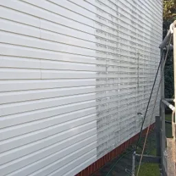 Boczna ściana domu z sidingu, w połowie umyta, ukazująca różnicę między czystą a zabrudzoną powierzchnią. W tle widoczny fragment rusztowania i zieleń.