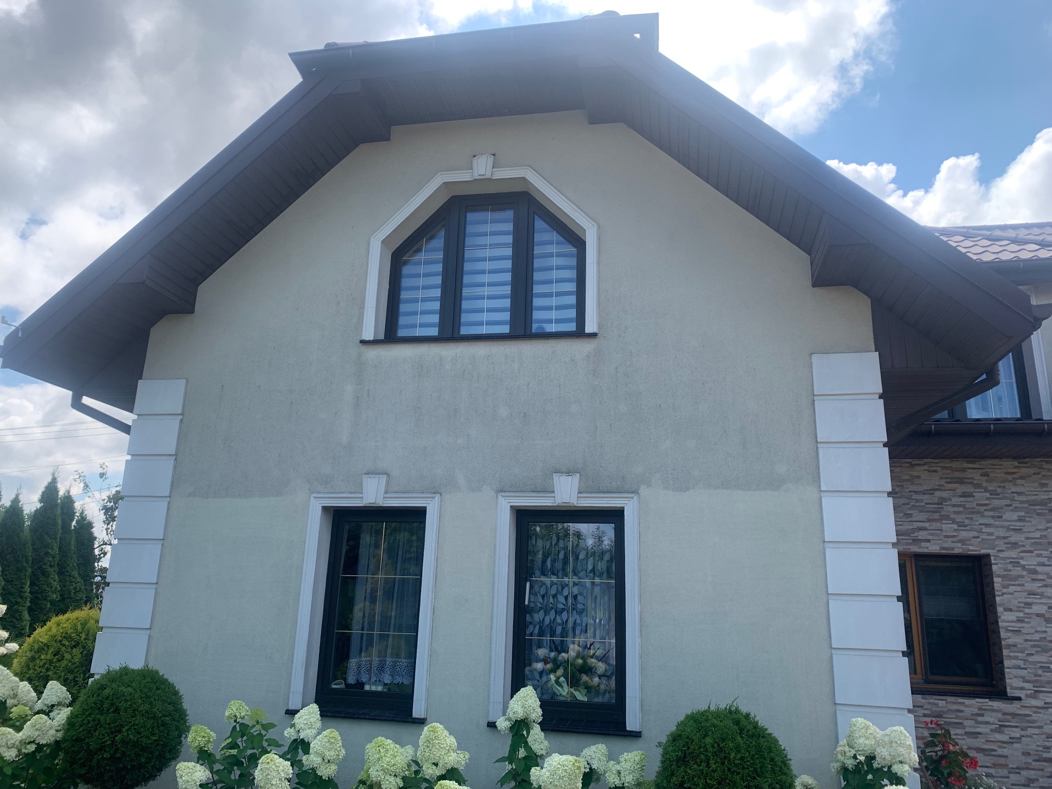 Dwukondygnacyjny dom z szarą elewacją, brązowym dachem i białymi detalami architektonicznymi, otoczony kwitnącymi hortensjami.
