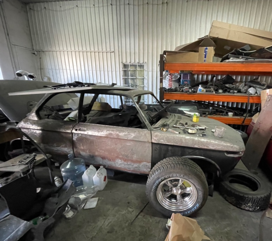 BMW 2002 w trakcie renowacji w warsztacie, z widocznymi śladami korozji i częściowo zdemontowanym nadwoziem, narzędzia leżące na masce, wnętrze częściowo odsłonięte, na pierwszym planie nowe koło...