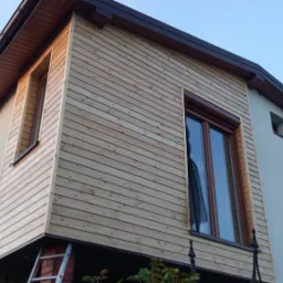 Fragment elewacji domu z nową, jasną, drewnianą okładziną, widoczne okna z brązowymi ramami i fragment drabiny.