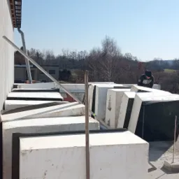 Prace przygotowawcze do ocieplenia dachu: Ułożone bloczki styropianowe z grafitowym wypełnieniem na dachu budynku, widoczny pracownik w bluzie z kapturem oraz krajobraz w tle.