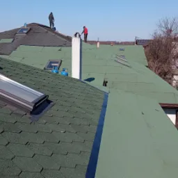 Prace dekarskie w Bytomiu: montaż okna dachowego na dachu pokrytym zielonym gontem bitumicznym, widoczne butle z gazem i dekarze na sąsiednim, częściowo wykończonym dachu.