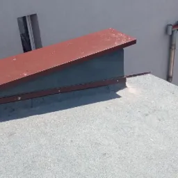 Fragment płaskiego dachu pokrytego papą z widocznym kominem obłożonym blachą w kolorze brązowym, w tle budynek mieszkalny i fragment ogrodu.