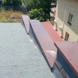 Widok z góry na płaski dach pokryty papą z zamontowanymi brązowymi obróbkami blacharskimi, w tle budynek z klimatyzatorami i drzewa.