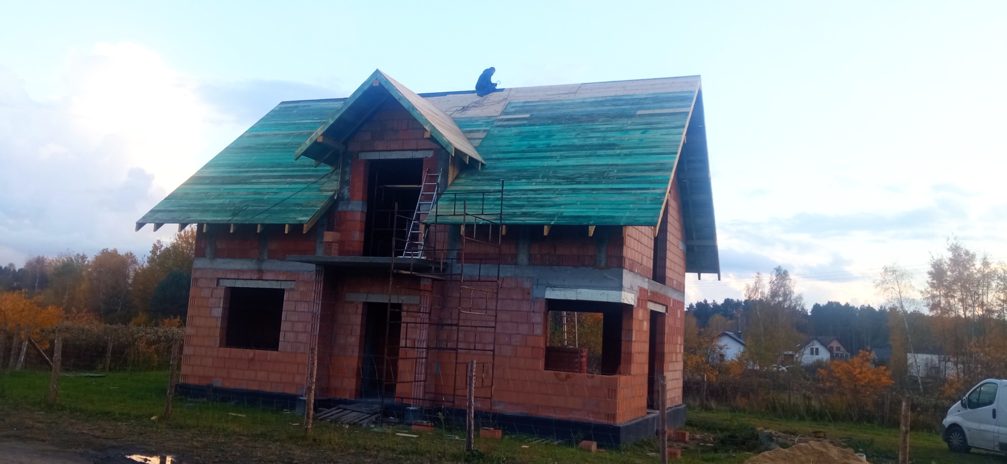 Surowy dom w trakcie budowy z częściowo ułożonym, zielonym deskowaniem dachu i pracującym na nim dekarzem, widoczne rusztowania i drabina.