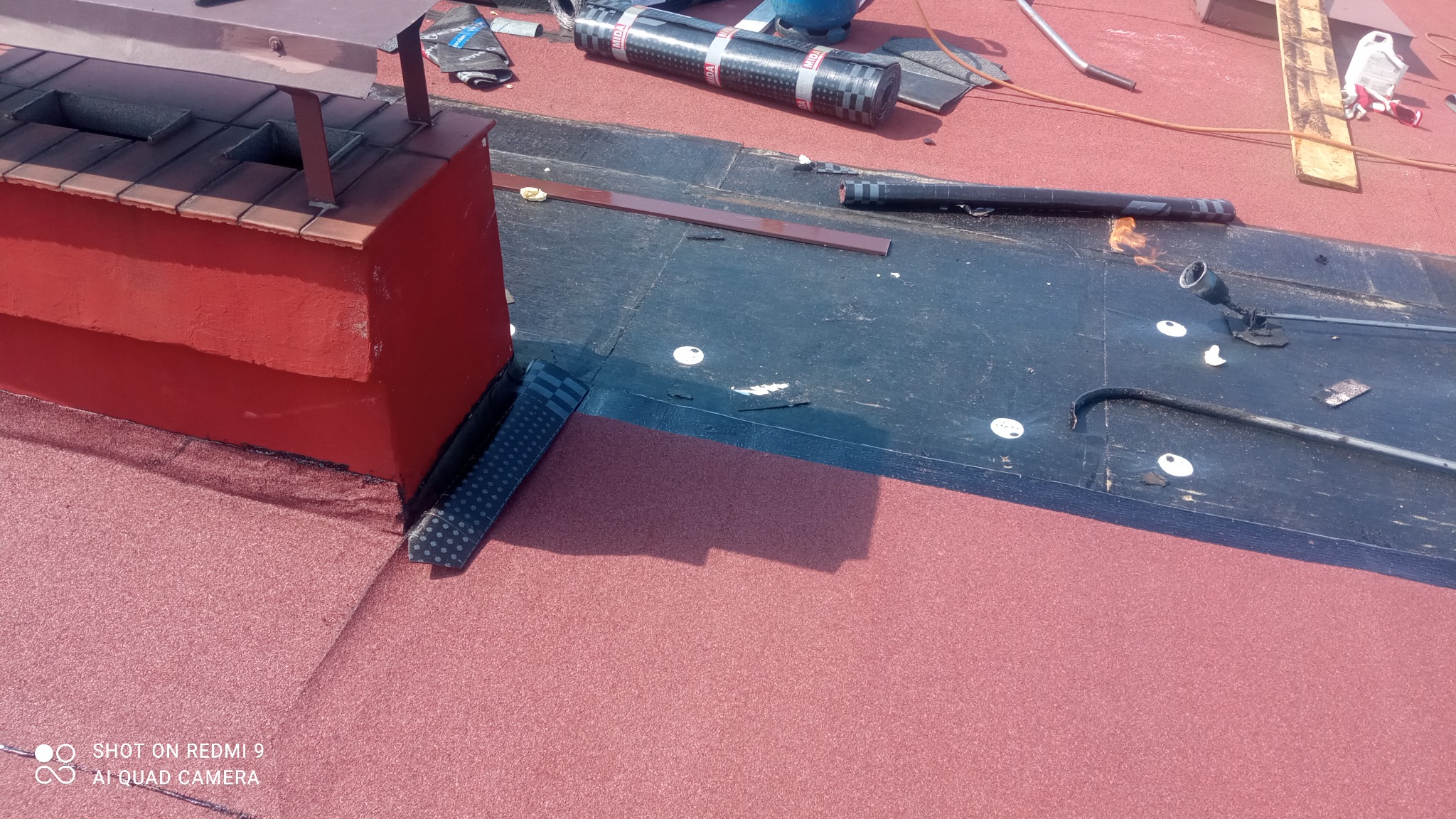 Nowa, czerwona papa na dachu z kominem w trakcie prac, widoczne narzędzia i materiały izolacyjne.