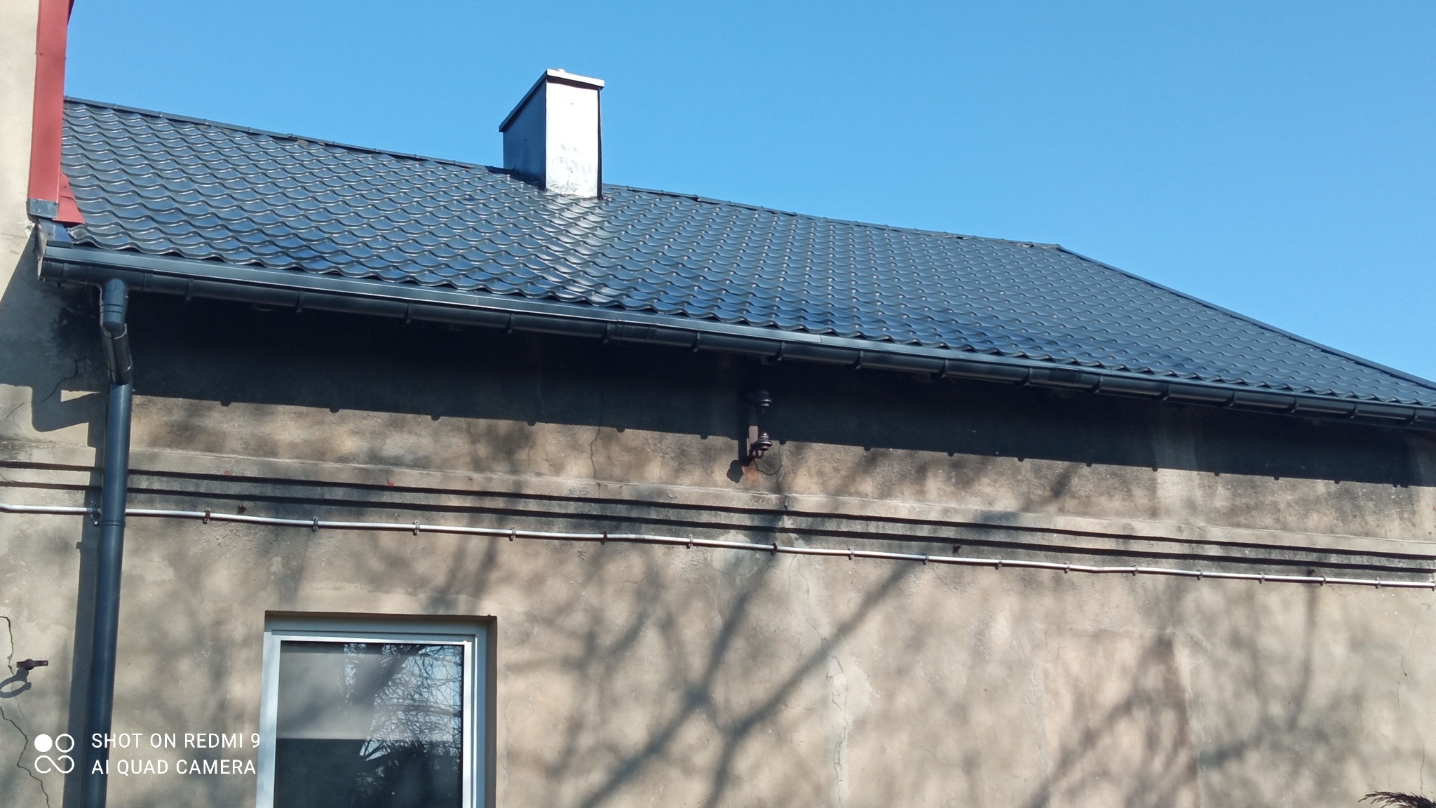 Nowy, ciemnoszary dach z blachy trapezowej z kominem i rynnami, widoczny fragment elewacji z oknem i instalacjami.
