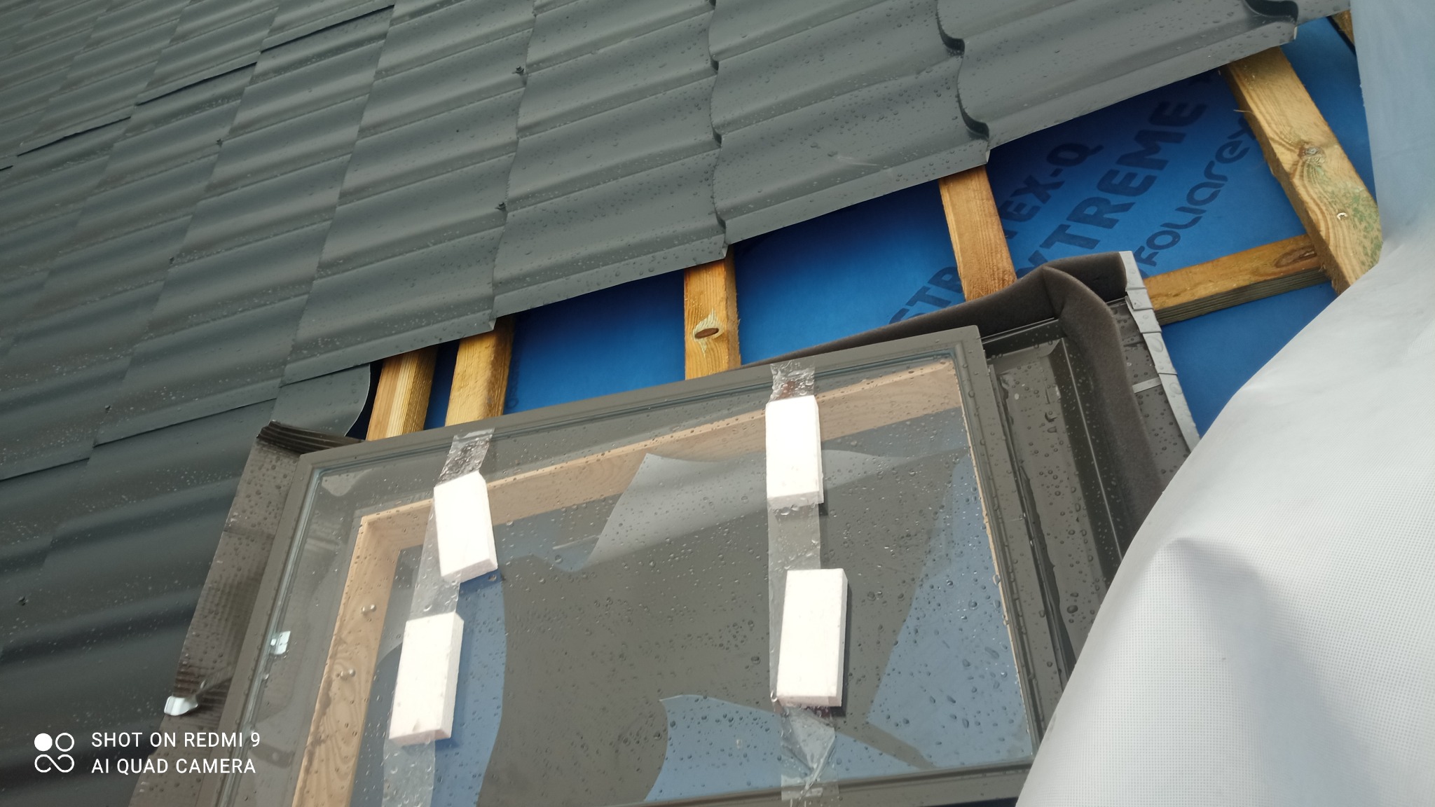 Montaż okna dachowego na tle połaci z blachy trapezowej, widoczna membrana dachowa i drewniana konstrukcja więźby, krople deszczu na powierzchni.