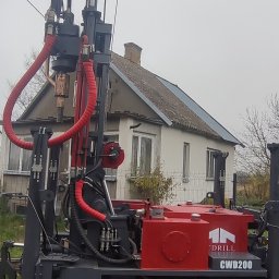 Dobry Wiertnik - Wiertnica gąsienicowa CWD200 na tle domu jednorodzinnego, widoczne czerwone przewody hydrauliczne i logo firmy wiertniczej.