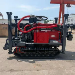 Wiertnica JCDRILL CWD200 na gąsienicach z czerwonymi przewodami hydraulicznymi na placu budowy, widoczny dźwig w tle.