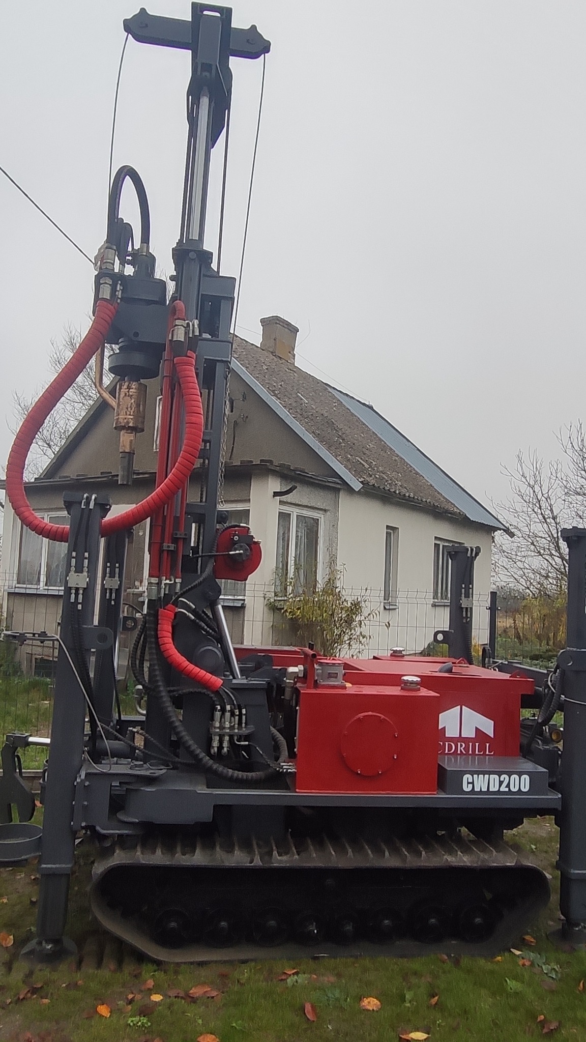 Wiertnica gąsienicowa CWD200 na tle domu jednorodzinnego, widoczne czerwone przewody hydrauliczne i logo firmy wiertniczej.