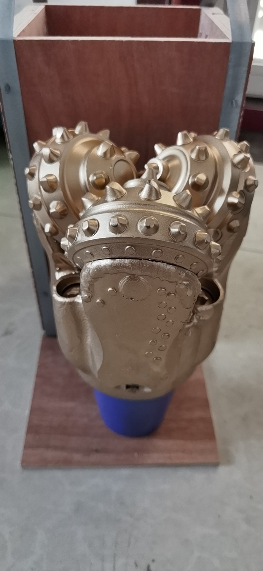 Złota głowica wiertnicza z trzema obrotowymi elementami tnącymi, umieszczona na niebieskiej podstawie na drewnianej powierzchni. Widoczne detale konstrukcyjne i ślady użytkowania.