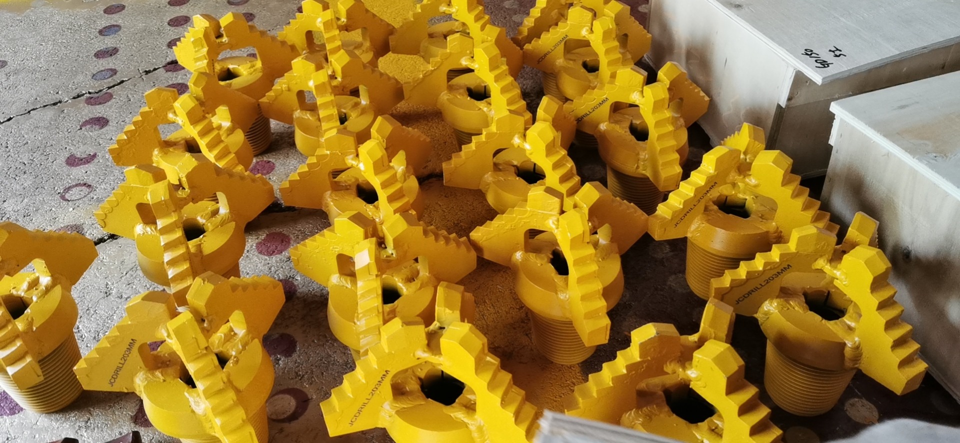 Żółte wiertła do studni z gwintem i oznaczeniem JCDRILL203MM ułożone na podłodze, obok drewniane skrzynie.