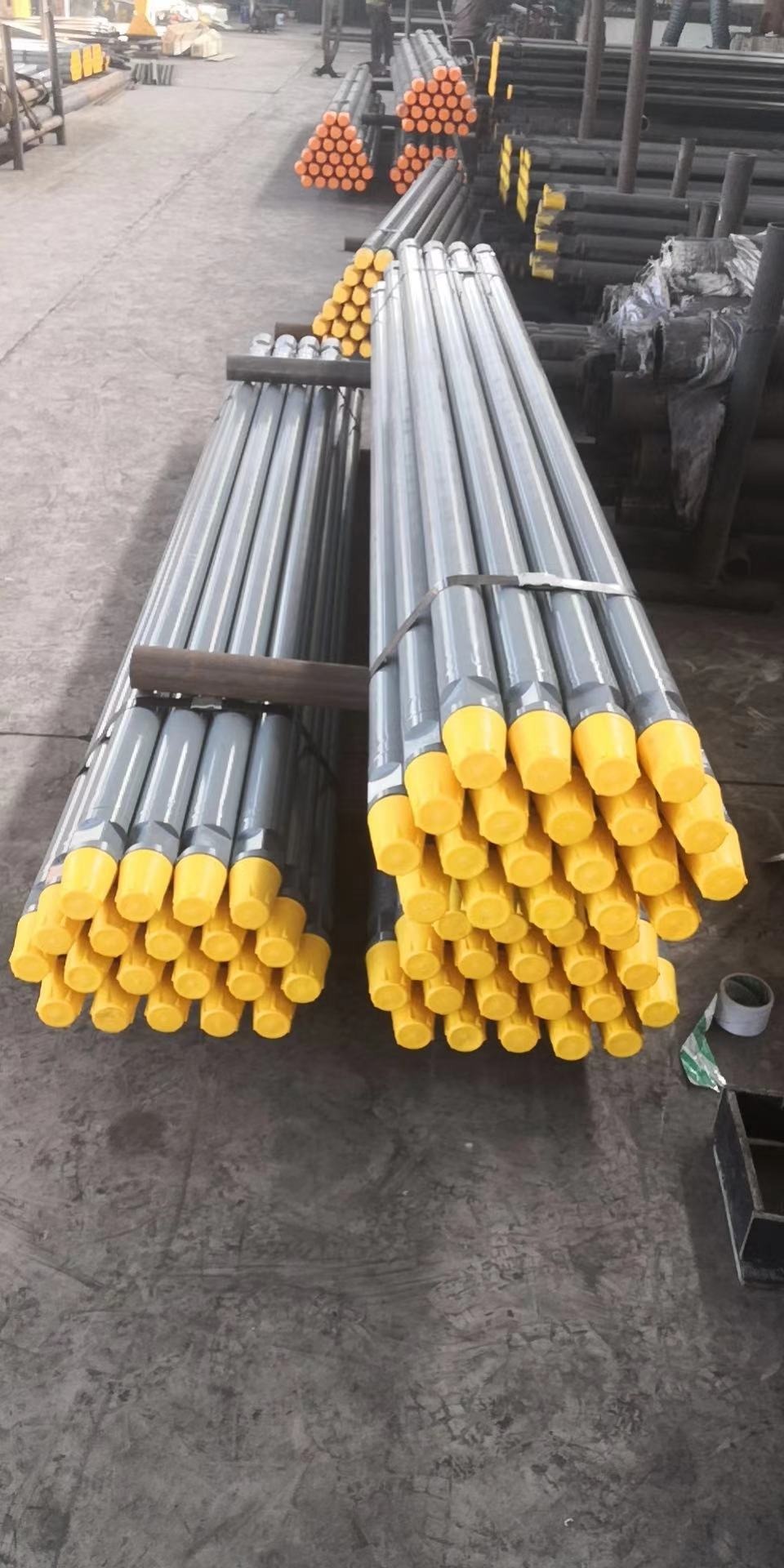 Pakiety szarych rur wiertniczych z żółtymi zaślepkami zabezpieczającymi, ułożone na betonowej podłodze w magazynie.