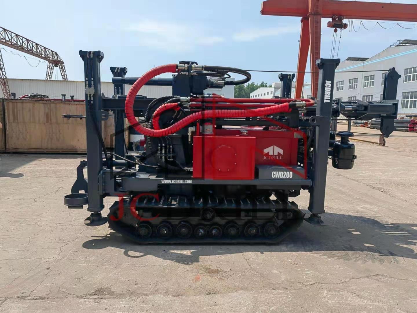 Wiertnica JCDRILL CWD200 na gąsienicach z czerwonymi przewodami hydraulicznymi na placu budowy, widoczny dźwig w tle.
