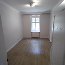 Sas-bud usługi ogólnobudowlane Wojciech Sas - Jasne, puste pomieszczenie po remoncie, z panelami podłogowymi w odcieniach drewna, pomalowanymi na biało ścianami i grzejnikiem pod oknem.