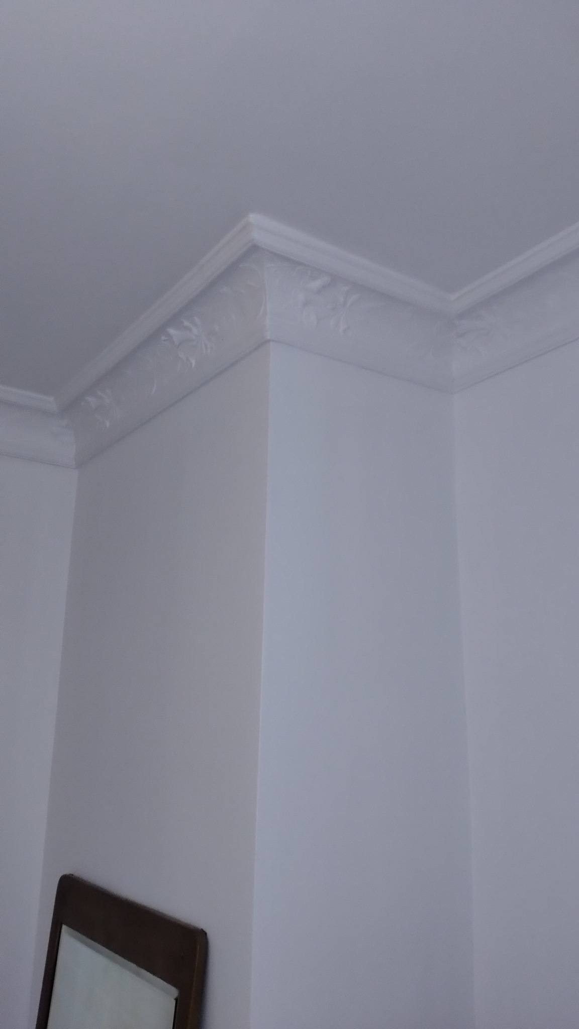 Pomieszczenie po malowaniu na biało, widoczna dekoracyjna listwa przysufitowa z motywem roślinnym na styku ścian i sufitu, fragment ramy lustra w rogu.