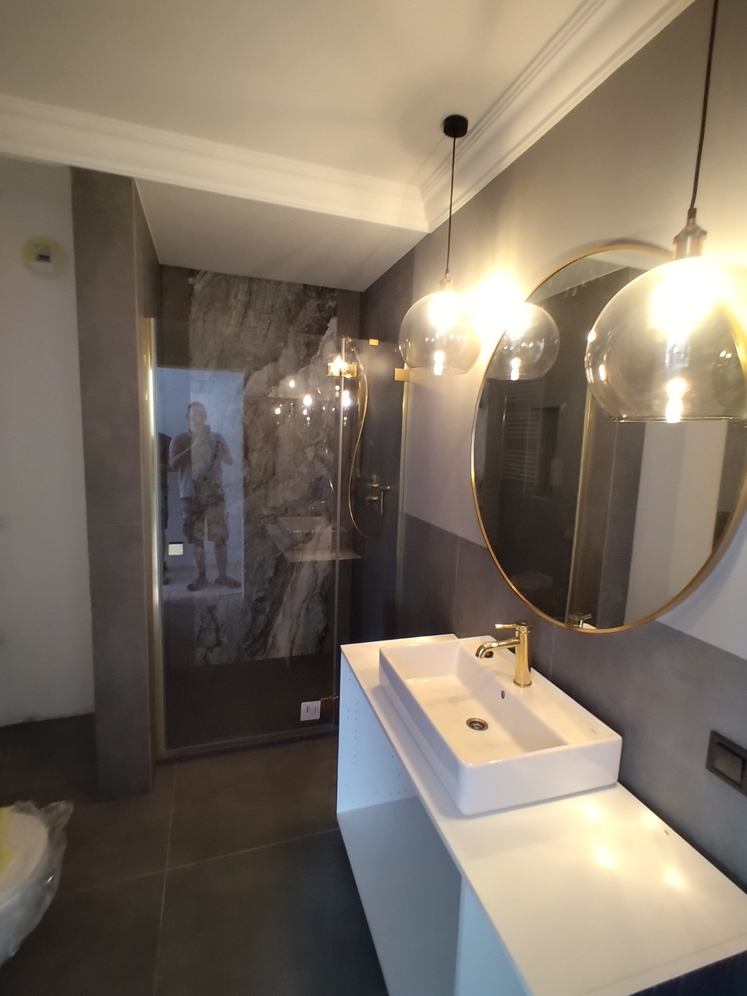 Nowoczesna łazienka z marmurową ścianą w kabinie prysznicowej, okrągłym lustrem w złotej ramie, białą umywalką z baterią w kolorze złota i wiszącymi lampami z przezroczystego szkła.