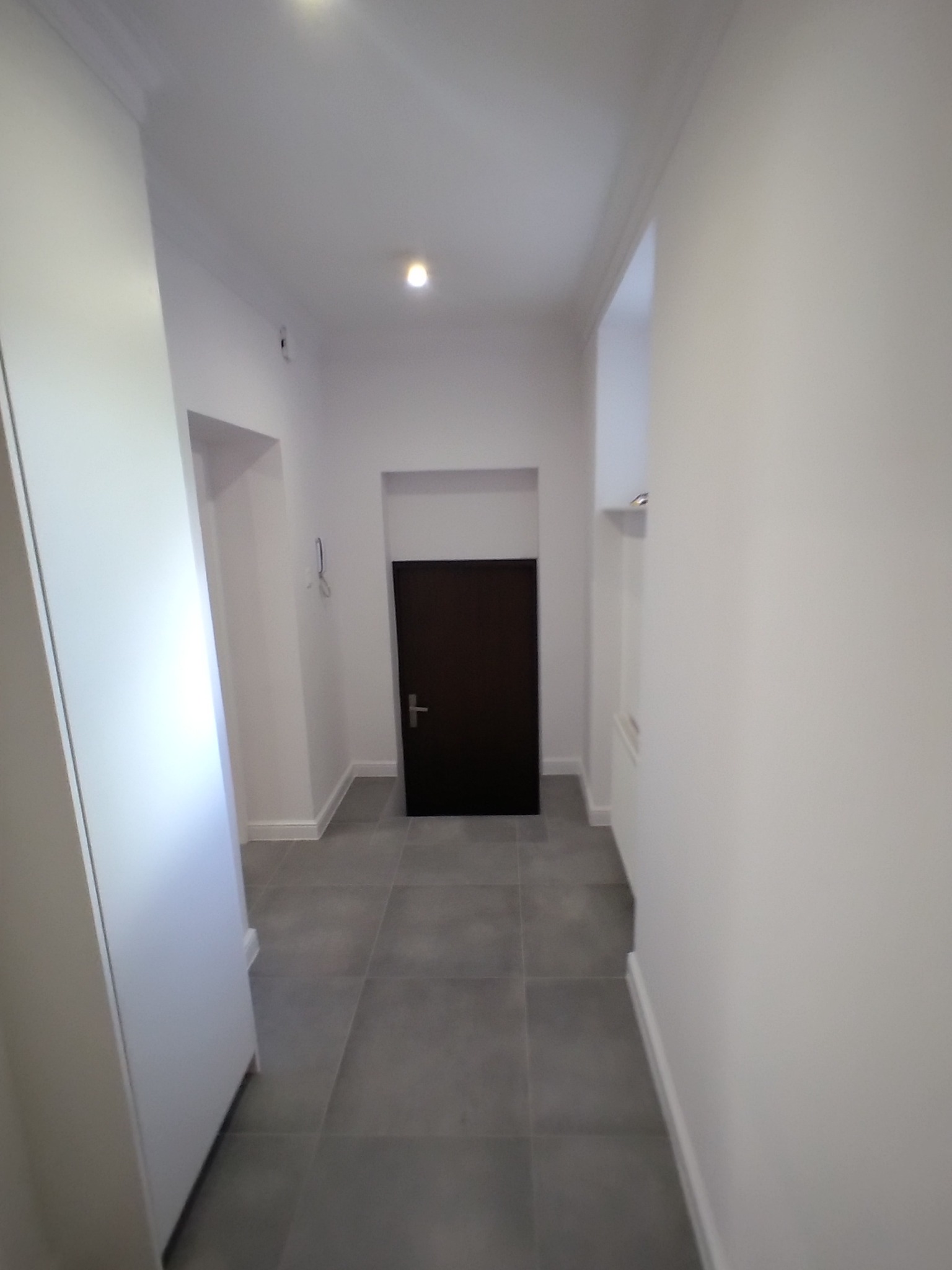 Wnętrze korytarza po remoncie w Poznaniu, z szarymi płytkami podłogowymi i świeżo pomalowanymi na biało ścianami, widok na ciemne drzwi na końcu korytarza.