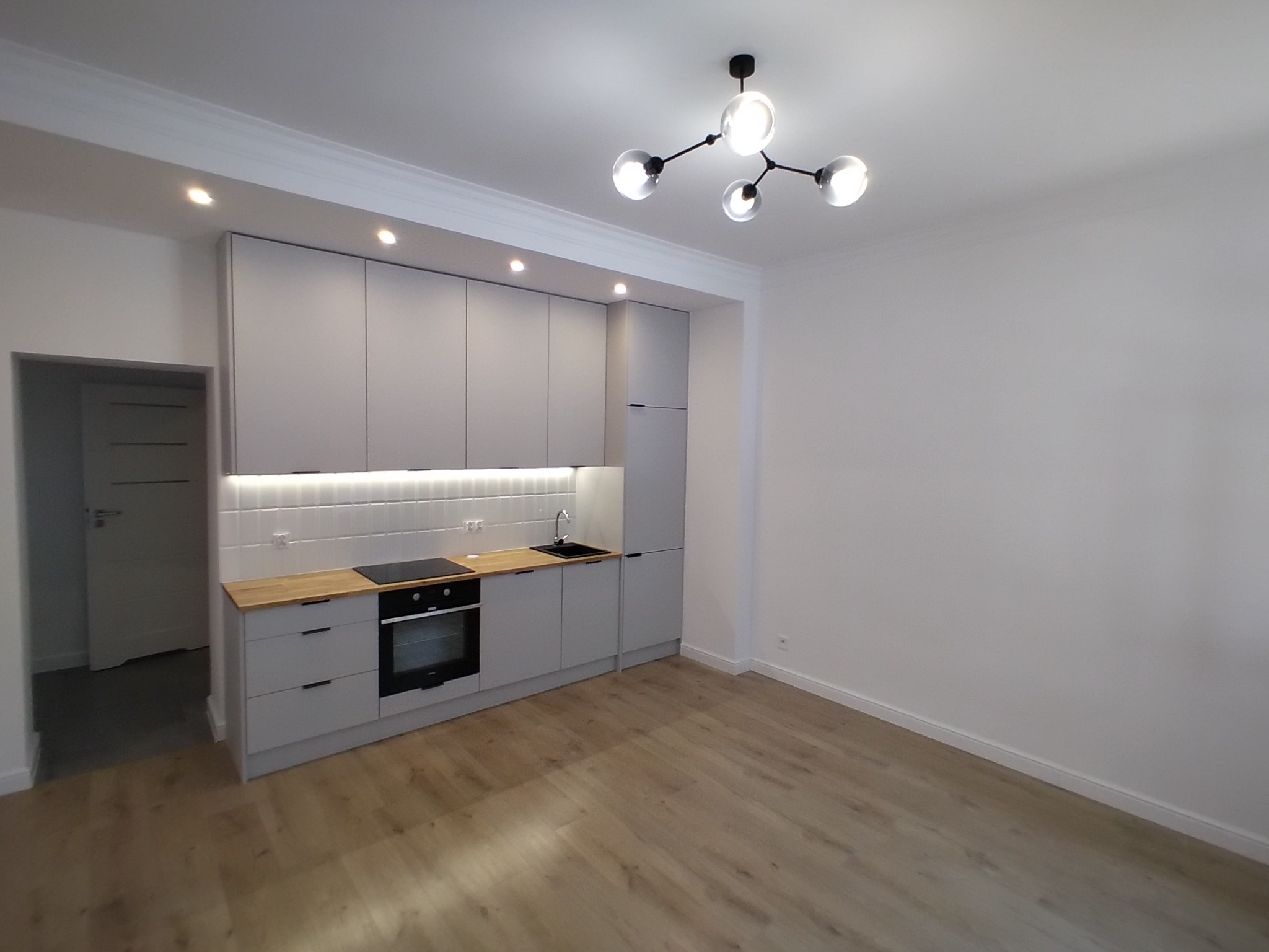 Szare, nowoczesne meble kuchenne z drewnianym blatem, białe płytki na ścianie, parkiet na podłodze i oryginalna lampa sufitowa w nowym mieszkaniu.