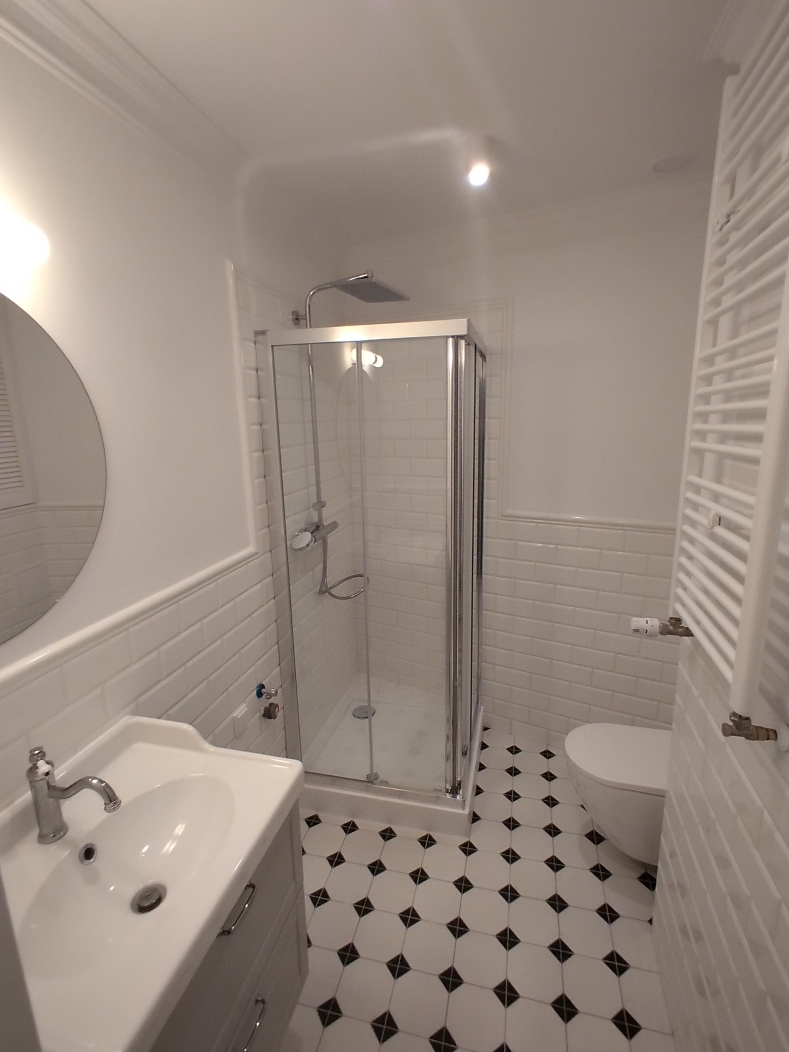 Nowoczesna łazienka z kabiną prysznicową, umywalką na szafce i grzejnikiem drabinkowym. Białe płytki imitujące cegłę na ścianach i czarno-biała podłoga w romby.