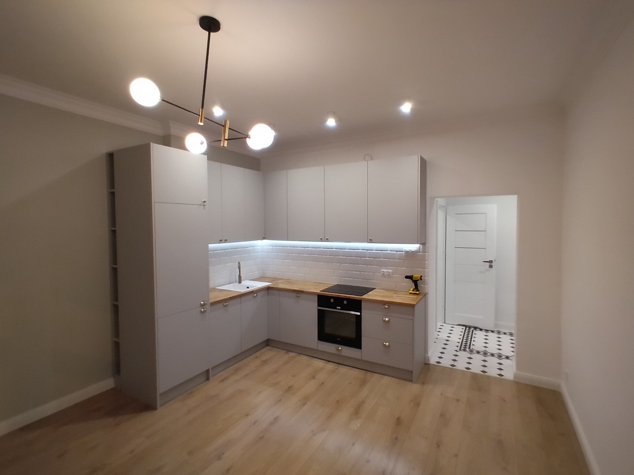 Nowoczesna, szara kuchnia z drewnianym blatem i oświetleniem LED, widok na przedpokój z wzorzystą podłogą, w tle wiertarka akumulatorowa na blacie.