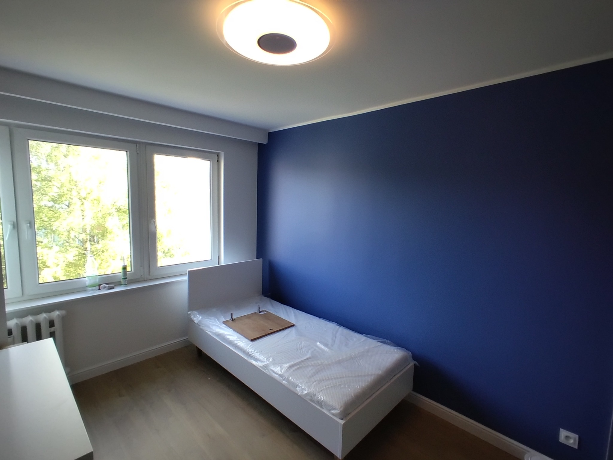 Pokój po malowaniu ścian na ciemny niebieski kolor, z białym łóżkiem zabezpieczonym folią malarską i fragmentem drewnianej deski na materacu, widok na okno z drzewami na zewnątrz.