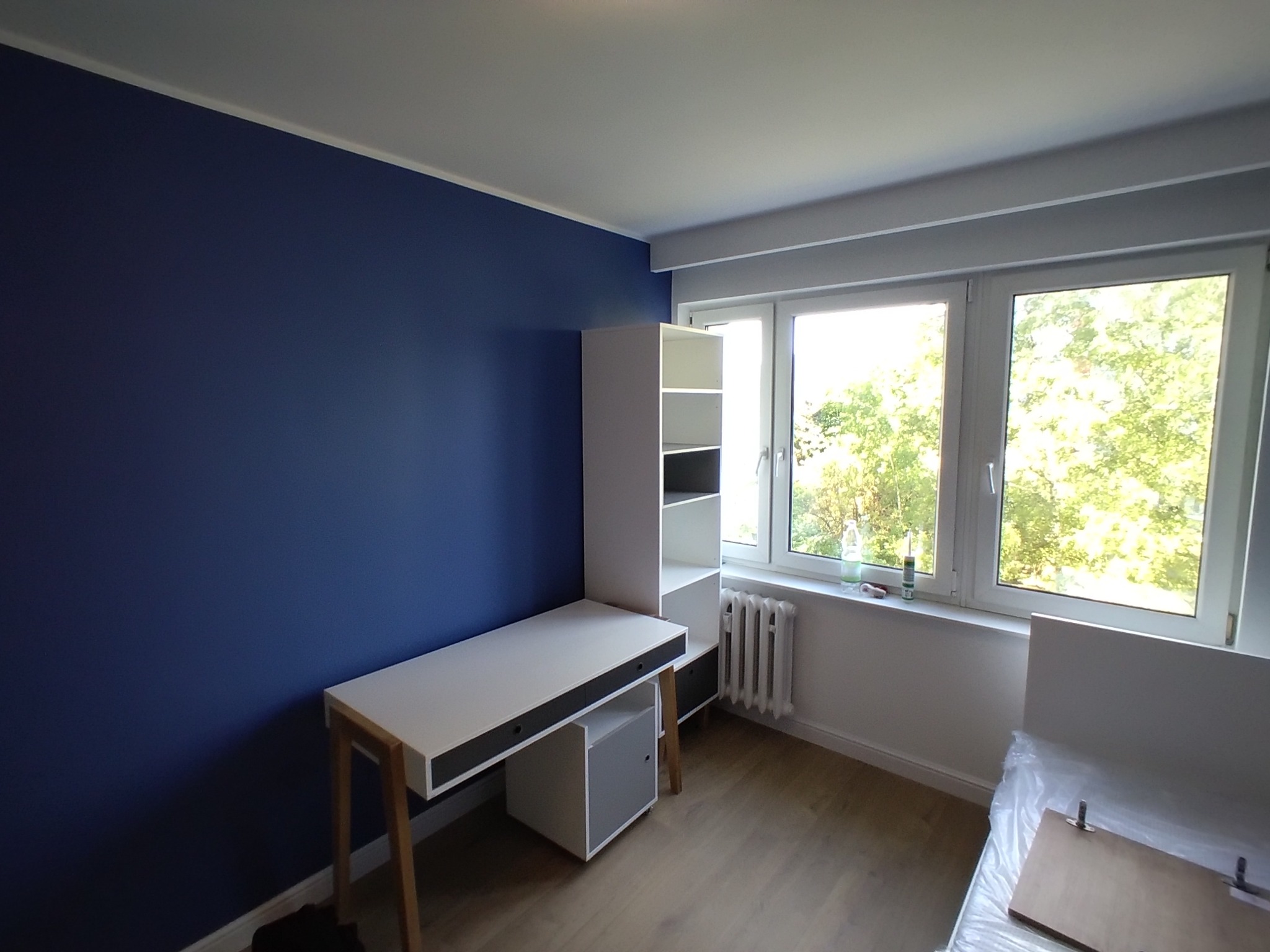Ściana pomalowana na granatowo w pokoju z białymi meblami, w tym biurko z regałem i grzejnik pod oknem.