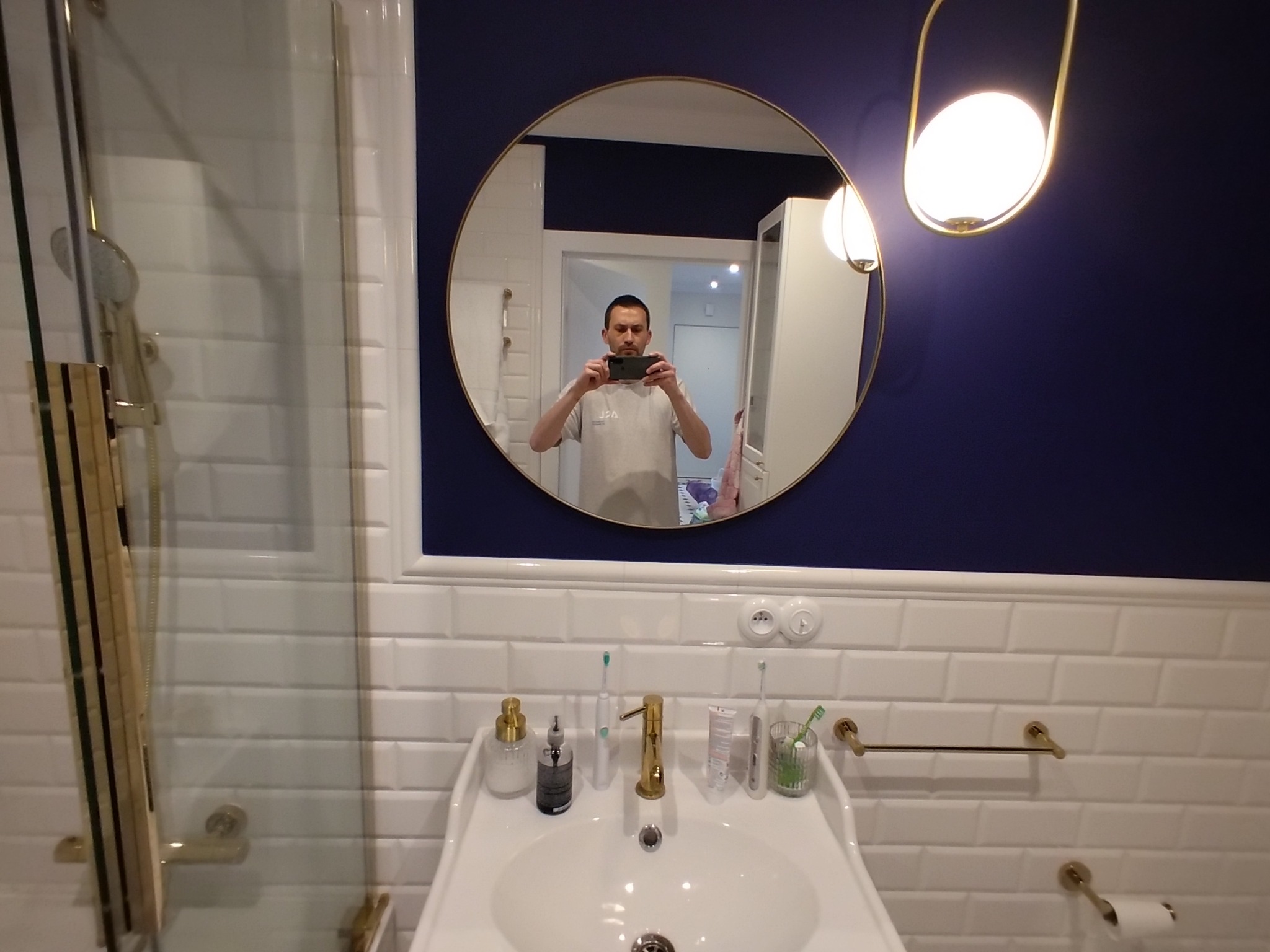 Nowoczesna łazienka z granatową ścianą, białymi płytkami metra, okrągłym lustrem i złotymi akcentami, widoczne odbicie osoby fotografującej.