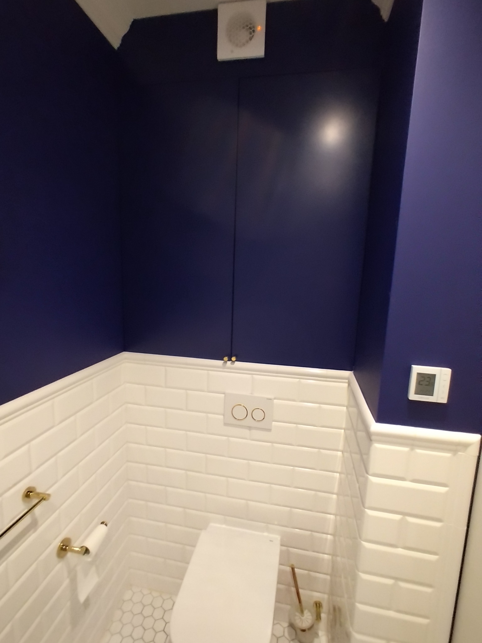 Niebieska ściana w toalecie z białymi płytkami typu metro, złotymi akcentami i ukrytym schowkiem nad spłuczką.